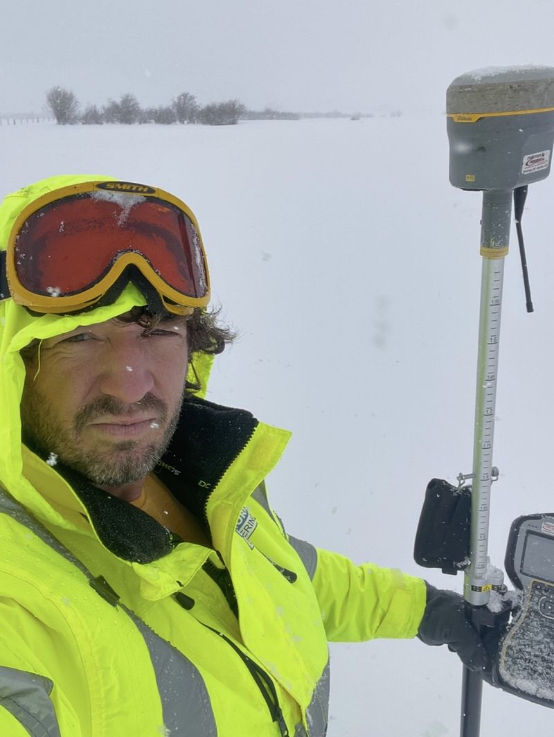 Jeremiah in a hi-vis jacket and goggles holding a GPS rover and data collector in a snowstorm