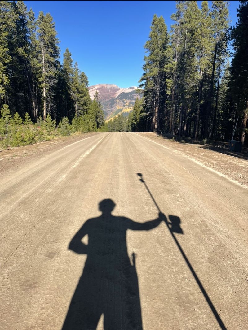 Shadow of a surveyor holding a GPS rover pole on a dirt road through pine forest, with a colorful mountain peak beyond